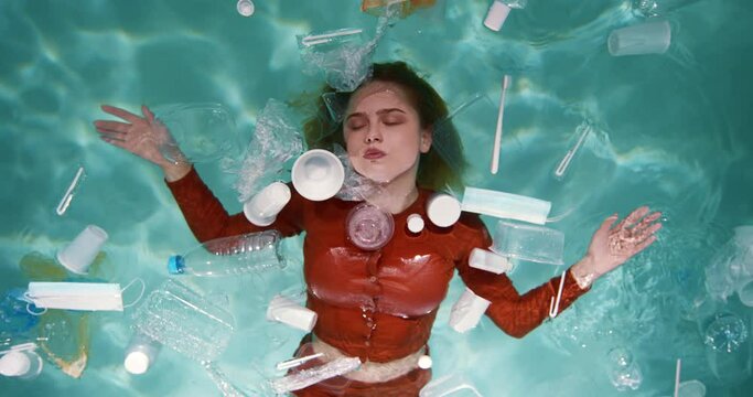Cinematic top view, beautiful woman lying on water surface surrounded by various plastic packaging waste slow motion.