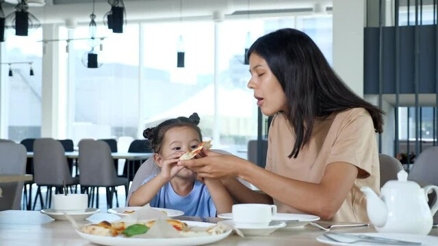 Mother and daughter eat pizza. Asian woman mom and toddler girl with space buns eat delicious snack playing at wooden table in local cafe close view