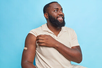 Horizontal shot of dark skinned bearded man happy to get vaccinated shows arm after receiving...