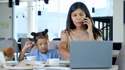 Hungry Daughter asks Mother to feed himself in restaurant. Asian businesswoman mom gives pizza slice to toddler child sitting at table with tea and grey laptop close-up - Powered by Adobe