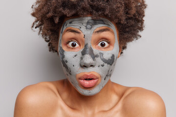 Surprised Afro American woman with curly hair stares impressed applies clay facial mask takes care of dry complexion gasps from wonder undergoes beauty treatments isolated over white background.
