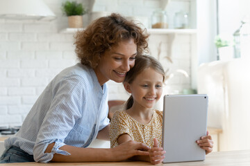 Smiling little girl with mother having fun with tablet together sitting at table in kitchen, happy mom and adorable kid girl looking at modern device screen, chatting online making video call