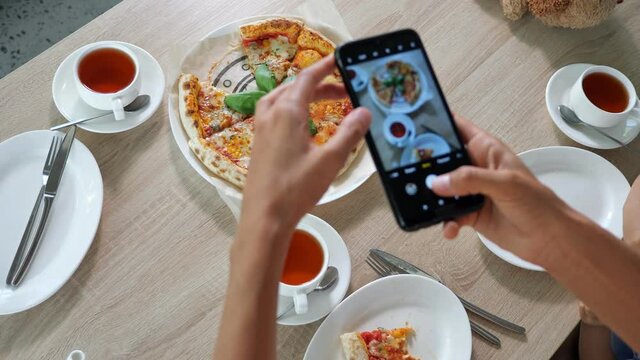 Lady films food. Young woman holds contemporary black smartphone and makes photo of delicious pizza and tea on wooden cafe table close view