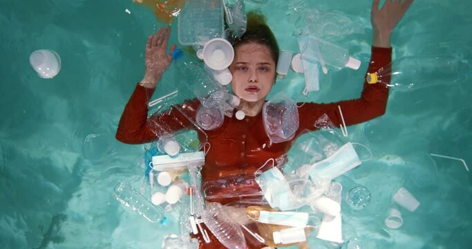 CARELESS USE OF PLASTIC. Cinematic top view, young beautiful woman on water surface among packaging waste slow motion.