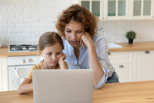 Focused Mother With Little Daughter Looking At Laptop Screen Together Studying Or Watching Webinar Sitting At Table In Kitchen, Pensive Mom And Preschool Girl Kid Pondering Problem With Device