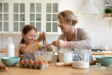 Smiling little girl with mature grandmother baking sitting at wooden table in modern kitchen...
