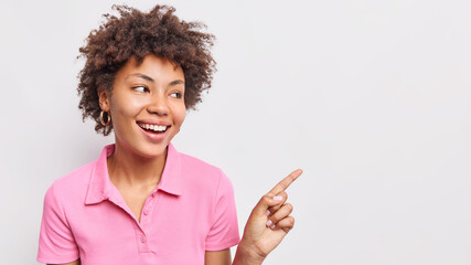 Photo of smiling curly haired Afro American woman points index finger right shows discount sale banner wears casual pink t shirt isolated over white background demonstrates cool awesome product