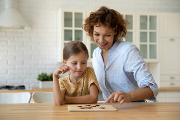 Smiling mother and little daughter playing logic strategy wooden board game at kitchen table...