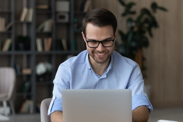 Working on laptop. Smiling young male worker wear glasses sit at desk use computer type document...