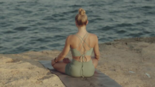 Young light hair woman doing meditation while practicing yoga overlooking mediterranean sea at sunset or sunrise. Mental and physical health. Calm meditative ambient. Slow motion video footage.