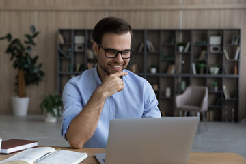 Research fellow. Smiling intelligent young man in glasses sit by laptop at modern scientific laboratory look on screen satisfied with job result. Male teacher having fun grading students papers on pc