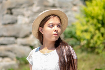 Portrait of a young girl of 17-20 years old in a white dress and a straw hat with long black hair against a stone wall and greenery.