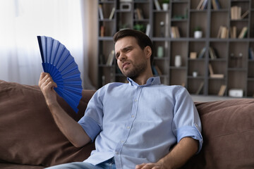 Too hot indoors. Exhausted male recline on sofa wave himself holding paper hand fan suffer of...