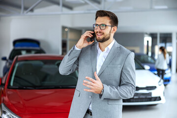 Smiling, friendly car seller standing in car salon and having a phone conversation with customer.