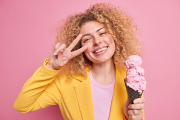Positive beautiful European woman with curly hair toothy smiles makes peace gesture over eye holds delicious big ice cream in waffle enjoys outdoor summer walk with friends wears formal outfit