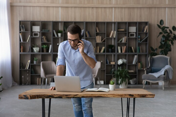 Keeping in touch. Young businessman talk on phone at workplace stand by laptop check information online. Busy male sales manager multitasking receive customer request by cell confirm order in web chat