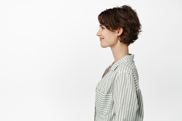 Profile portrait of beautiful smiling woman with short pixie hairstyle, looking left with happy smile, casual friendly face expression, standing in casual shirt over white background © Cookie Studio