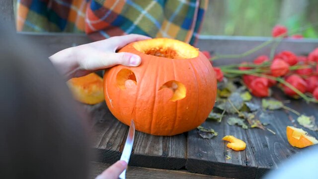 Hands carves a scary halloween pumpkin on a wooden bench in the garden. Making pumpkin decor for Halloween. Preparation for celebration.