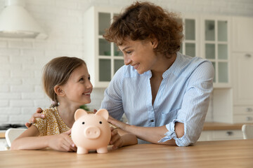 Happy mother and cute little girl holding pink piggy bank sitting at kitchen table, smiling mom...