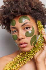 Spa female model with Afro curly hair looks seriously at camera has confident expression makes skin care treatments applies hydrogel patches on face holds wild plant undergoes beauty procedures