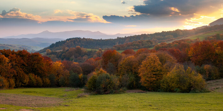 countryside mountain scenery at sunset. beautiful rural landscape in autumn. fields and meadow on rolling hills in evening light. trees in colorful foliage. ridge with high peak in the distance