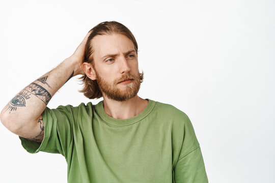Image of sassy handsome bearded guy with tattoos, hipster touching his long haircut, running fingers through hair and looking aside, standing against white background