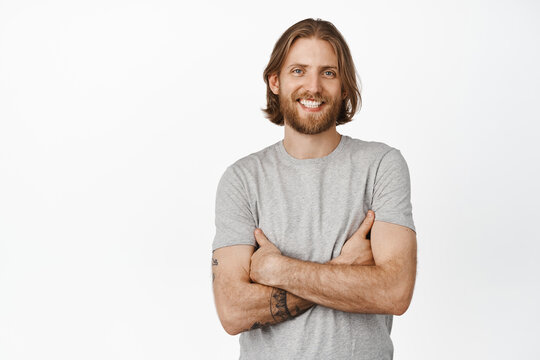 Image of handsome bearded guy with blond hairstyle, cross arms on chest, smiling white teeth, looking happy, standing in grey t-shirt against white background