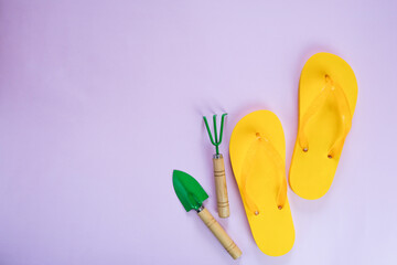 Gardening tools, green shovel and rake on yellow flip flops, on a pink or lilac background, close-up.
