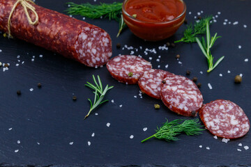 Salami sausage with ketchup and spices on black slate board