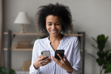 Smiling young African American woman user hold two smartphones synchronize data information on gadgets. Happy millennial mixed race female client use old new cellphones. Technology concept.