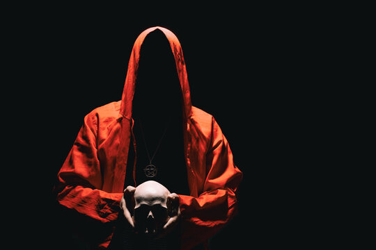 Man in red ritual hooded cloak holds a human skull in hands. Religious sects, satanism, occult, esoteric, concept. Copy space.
