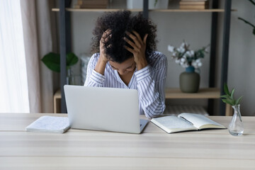 Unhappy young African American woman sit at desk at home office suffer from migraine or headache. Unhealthy mixed race female feel tired finish work deadline, lost motivation. Health problem concept.