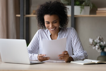 Happy young African American woman sit at desk at home office work online on computer with paperwork documents. Smiling biracial female read good news in paper correspondence. Success concept.