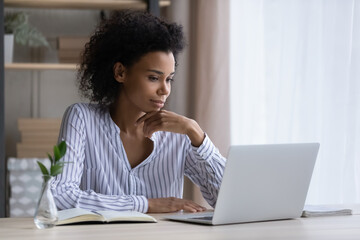 Happy pensive young African American woman sit at desk at home office look at laptop screen study or work online. Millennial ethnic female use computer browse internet on gadget. Technology concept. - Powered by Adobe