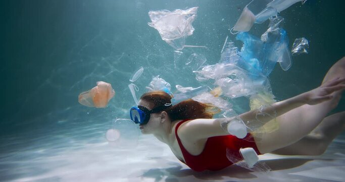 Concept of plastic pollution. Young female tourist swims through single use packaging waste under water slow motion.