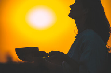 Silhouette of woman playing a Tibetan bowl at gorgeous sunset with sun disk orange gradient. Concept: peace, tranquility, zen, spirituality, sound healing