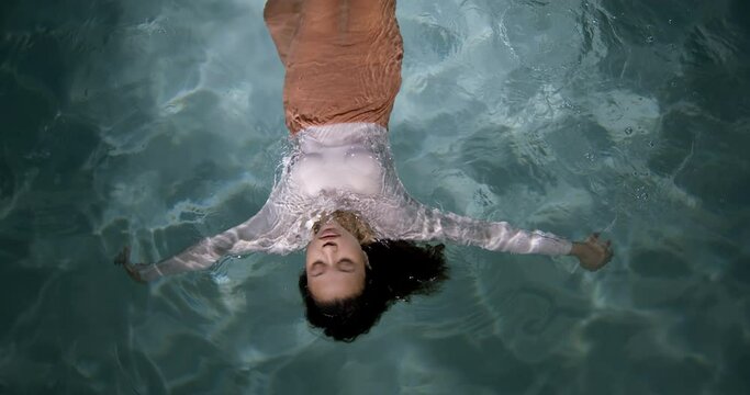 Depression Era. Cinematic Top View Of Young Beautiful Sad Mixed Race Woman Floating On Light Water Surface Slow Motion.