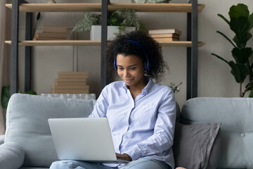 Young African American woman in headphones sit on sofa work online on laptop talk speak on video call. Happy biracial female in earphones have webcam digital virtual event on computer at home.