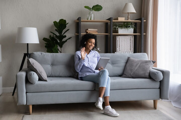 Smiling young African American woman sit on sofa at home wave greet talking speaking on video call on pad. Happy biracial female have webcam digital virtual event on tablet. Communication concept.