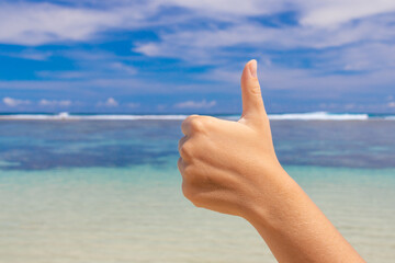 A female hand shows a thumbs up against a background of blue sea and sky. The concept of support, approval and good rest.