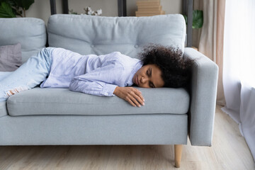 Exhausted young African American woman feel fatigue sleep or take nap on couch in living room....