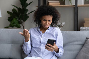 Unhappy young African American woman look at smartphone screen confused by error or mistake online on device. Upset biracial female use gadget feel distressed with bad negative news on cellphone. - obrazy, fototapety, plakaty