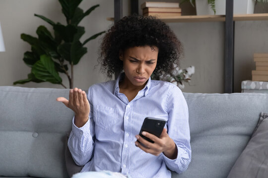 Unhappy Young African American Woman Look At Smartphone Screen Confused By Error Or Mistake Online On Device. Upset Biracial Female Use Gadget Feel Distressed With Bad Negative News On Cellphone.