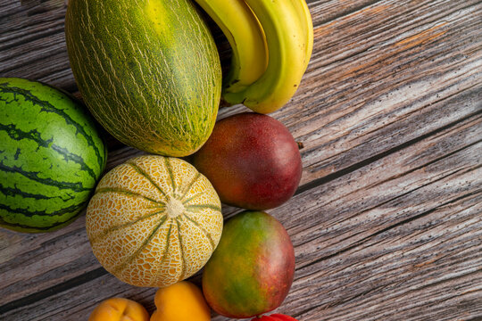 Top view image of ripe African mangoes, European melons and watermelons, canary bananas and yellow peaches on the wooden table.