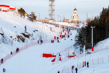 a ski slope for beginners with a lot of people on it and a fence, a chapel and trees on top of the slope. Russia - January 30, 2021