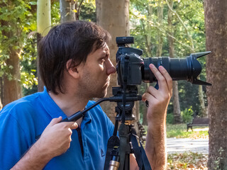 young white male photographer with blue t-shirt looks into the camera lens with a large lens mounted on a tripod, one hand adjusts focus, in the park on a sunny summer day