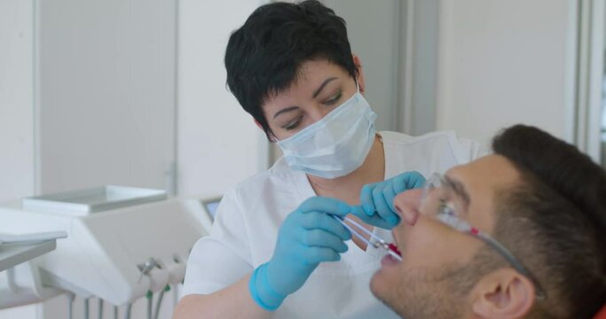 A female dentist in a face mask is holding in the male Arabian patient's mouth a dental articulating holder with paper and he is biting it. 