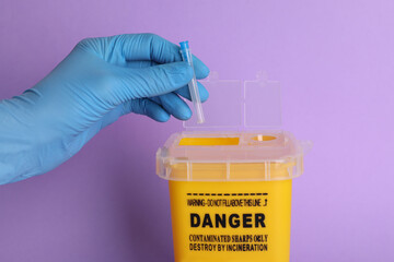 Doctor throwing used syringe needle into sharps container on violet background, closeup