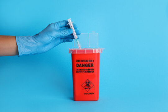 Doctor throwing used syringe into sharps container on light blue background, closeup