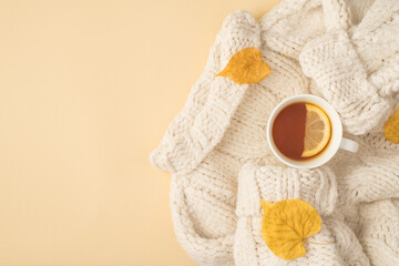 Top view photo of white sweater yellow autumn leaves and cup of tea with lemon on isolated beige background with copyspace